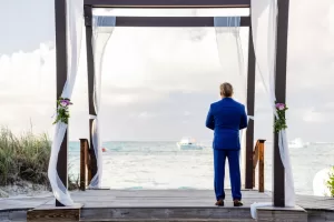 Professional Photographer A man in a blue suit stands under a wooden wedding arch draped with white fabric, gazing at the ocean with boats in the distance—a perfect moment for wedding photography on the sandy, grassy beach. www.maranonimagery.com