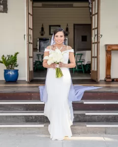 Professional Photographer A bride in a white gown holding a bouquet of white flowers stands smiling on steps in front of an open door, as if posing for elegant portraits, with a plant in a blue pot and wooden furniture visible in the background. www.maranonimagery.com