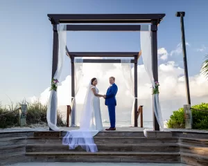 Professional Photographer A bride and groom stand holding hands under a wooden pergola with sheer white drapes, outdoors by the sea, sunlight and greenery all around—a perfect moment for stunning wedding photography. www.maranonimagery.com