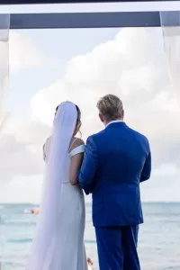 Professional Photographer A bride in a white dress and veil stands next to a groom in a blue suit, both facing the ocean under a canopy, with clouds and water in the background—perfect for timeless wedding photography and romantic portraits. www.maranonimagery.com