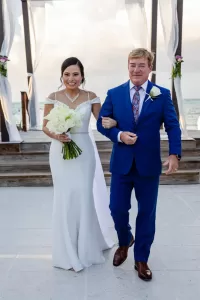 Professional Photographer A bride in a white gown holding a bouquet of white roses walks arm-in-arm with a man in a blue suit and floral tie. They are smiling as the photographer captures wedding photography portraits by a decorated arch with the ocean in the background. www.maranonimagery.com
