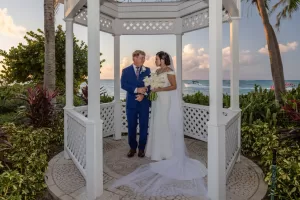 Professional Photographer A bride and groom stand together in a white gazebo surrounded by tropical plants, with the ocean and blue sky in the background. This romantic moment captures the essence of wedding photography, with the bride holding white flowers and the groom in a blue suit. www.maranonimagery.com