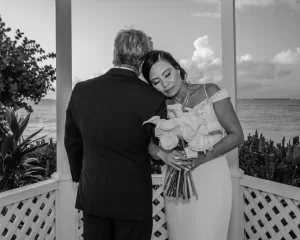 Professional Photographer A bride in a white dress rests her head on the groom’s shoulder, eyes closed, holding a bouquet of white flowers as a photographer captures their portrait in a gazebo by the ocean. The groom stands with his back to the camera. www.maranonimagery.com
