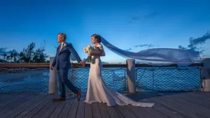 Professional Photographer A bride and groom walk hand in hand on a wooden pier at sunset, captured by a skilled photographer. The bride’s veil flows in the wind as she holds a bouquet, with the ocean and blue sky creating a perfect backdrop. www.maranonimagery.com