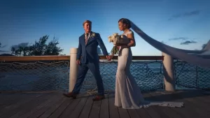 Professional Photographer A bride and groom walk hand in hand on a wooden pier at sunset. Perfect for wedding photography, the bride’s white dress and veil contrast beautifully with the groom’s blue suit against the ocean and trees in the background. www.maranonimagery.com