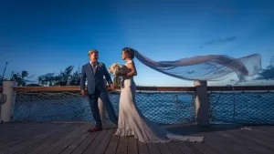 Professional Photographer A bride and groom stand on a wooden deck by the ocean at sunset. Capturing this magical moment, expert wedding photography highlights her flowing veil and bouquet, with clear skies and tree silhouettes completing the romantic scene. www.maranonimagery.com
