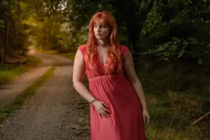 Professional Photographer A woman with long red hair in a pink dress stands on a gravel path in a wooded area, looking to the side with a thoughtful expression—an ideal setting for portraits captured by an experienced photographer amid lush greenery and trees. www.maranonimagery.com