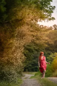 Professional Photographer A woman in a pink dress walks barefoot along a gravel path surrounded by lush green trees and foliage, as a photographer captures natural portraits in the soft sunlight filtering through the leaves. www.maranonimagery.com