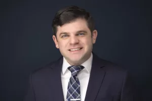 Professional Photographer A man in a dark suit, white shirt, and plaid tie smiles in front of a dark blue background—perfect for professional headshots or portraits. www.maranonimagery.com