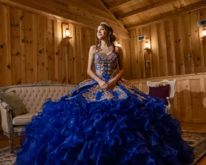 Professional Photographer A young woman in a blue and gold ball gown and tiara stands in a wooden room, smiling and posing elegantly near an ornate sofa, perfect for wedding photography. www.maranonimagery.com
