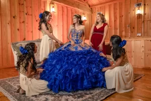 Professional Photographer A young woman in an elaborate royal blue and gold gown stands in the center of a wooden room, surrounded by four smiling women in matching cream and gold dresses, as a photographer captures the joyful moment they help arrange her dress. www.maranonimagery.com