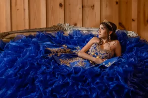 Professional Photographer A young woman in a blue and gold embroidered gown with a large ruffled skirt reclines on an ornate sofa, wearing a tiara, in a wooden-paneled room—an elegant scene perfect for portraits by a skilled photographer. www.maranonimagery.com