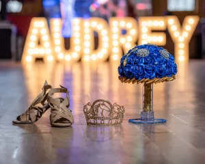 Professional Photographer A pair of beige dance shoes, a gold crown, and a blue rose bouquet are displayed on a wooden floor. In the background, large lighted letters spell AUDREY—perfect props for any creative photographer capturing unique headshots. www.maranonimagery.com