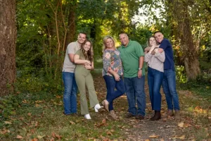 Professional Photographer Three couples stand close together on a leaf-strewn path in a wooded area, smiling and posing for the photographer. Sunlight filters through the green trees, creating a perfect setting for natural wedding photography portraits. www.maranonimagery.com