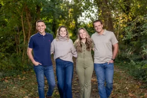 Professional Photographer Four smiling adults stand arm in arm on a wooded path, dressed in casual clothes. Sunlight filters through green trees as a photographer captures portraits in this warm, cheerful atmosphere. www.maranonimagery.com
