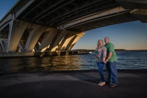 Professional Photographer A smiling couple stands hand in hand near the waterfront under a large bridge, with sunlight casting shadows and water and sky visible in the background—an ideal scene for wedding photography by an experienced photographer. www.maranonimagery.com