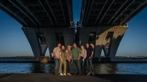 Professional Photographer Five people stand together under a large concrete bridge by the water at sunset, casting long shadows. Smiling and posing closely as a group, they create a memorable moment perfect for portraits by a talented photographer. www.maranonimagery.com