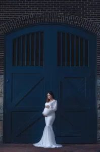 Professional Photographer A pregnant woman in a long, white dress stands in front of large, dark blue barn doors. Captured by a skilled photographer, she gently holds her belly and looks down with a serene expression. www.maranonimagery.com