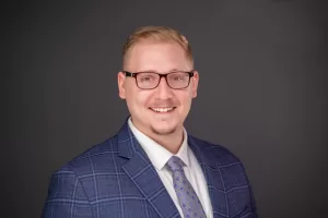 Professional Photographer A man with short blond hair and glasses smiles, wearing a blue plaid suit jacket, white shirt, and patterned tie, against a plain dark background—an ideal subject for portrait photography. www.maranonimagery.com