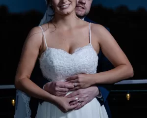 Professional Photographer A bride in a white dress and veil is smiling as her groom stands closely behind her, both holding hands around her waist at night with string lights in the background—a magical moment perfect for romantic portraits. www.maranonimagery.com