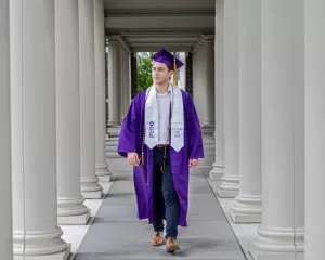 Professional Photographer A young man in a purple graduation cap and gown walks confidently between white columns. Captured by a talented photographer, he wears a white honor stole and cords, with Class of 2024 visible, and looks ahead with a slight smile. www.maranonimagery.com