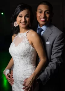 Professional Photographer A smiling couple poses together for wedding photography, with the woman in a white lace wedding dress and the man in a gray suit. They embrace, standing closely, and look directly at the camera in a dark, indoor setting. www.maranonimagery.com