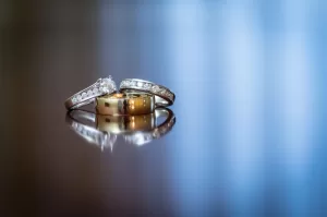 Professional Photographer Three rings, including a gold wedding band and two diamond-studded rings, rest together on a reflective surface with a soft blue background—an elegant moment captured by a skilled wedding photography professional. www.maranonimagery.com
