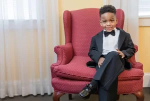 Professional Photographer A young boy in a black tuxedo and bow tie sits smiling on a red patterned armchair in a warmly lit room with cream-colored walls and white curtains—an ideal moment for charming wedding photography or headshots. www.maranonimagery.com