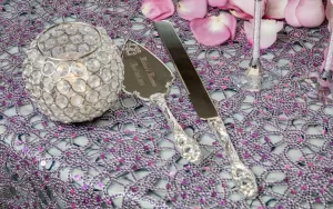 Professional Photographer A jeweled cake knife and server set rests on a purple beaded tablecloth, ready for the photographer to capture every detail—next to a round crystal candle holder and scattered pink rose petals, perfect for wedding photography. www.maranonimagery.com