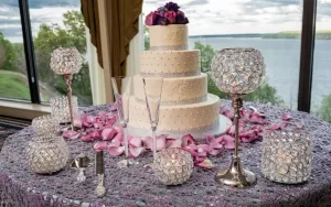 Professional Photographer A three-tiered white wedding cake with purple flowers sits on a sparkly purple cloth, surrounded by crystal candle holders, rose petals, and champagne flutes—perfect for capturing stunning wedding photography by the window overlooking the lake. www.maranonimagery.com