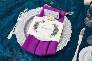 Professional Photographer A wedding place setting with a silver charger plate, purple napkin, and a card labeled Groom with floral design, set on a teal tablecloth alongside silverware and wine glasses—perfect for elegant wedding photography. www.maranonimagery.com