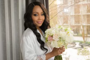 Professional Photographer A woman in a white robe stands indoors by a window, holding a bouquet of white and pale pink flowers, smiling softly with loose wavy hair. With trees and buildings outside, the scene radiates classic wedding photography elegance. www.maranonimagery.com