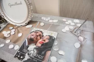 Professional Photographer A wedding guestbook table with a framed “Guestbook” sign, pen, scattered white and pink rose petals, and a personalized guestbook featuring a Wedding Photography portrait of the couple labeled “Dina & Andy, August 16th, 2018.”. www.maranonimagery.com