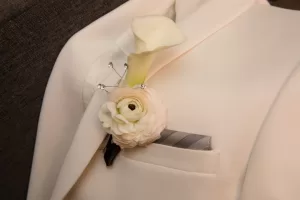 Professional Photographer A close-up of a white suit jacket with a grey pocket square and a boutonniere featuring a white calla lily and cream ranunculus, accented with decorative pins—perfect for wedding photography or elegant portraits. www.maranonimagery.com
