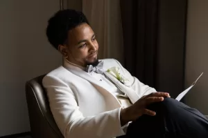 Professional Photographer A man in a white tuxedo jacket with a boutonniere sits in a chair, smiling thoughtfully while holding a piece of paper, as soft indoor lighting highlights his face—perfectly capturing the skill of a talented photographer known for stunning headshots. www.maranonimagery.com