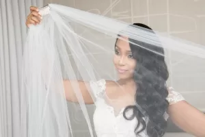 Professional Photographer A bride with long, wavy hair in a lacy white dress holds up a sheer veil and smiles softly. Perfect for wedding photography, the neutral background and soft lighting create an elegant portrait. www.maranonimagery.com