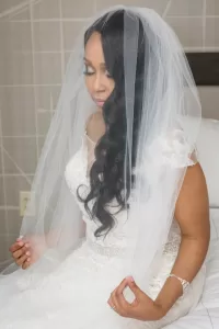Professional Photographer A bride in a white lace wedding dress and veil sits on a bed, gently holding the edge of her veil with both hands, looking down with a serene expression—an intimate moment captured through elegant wedding photography. www.maranonimagery.com