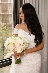 Professional Photographer A bride in an off-shoulder lace wedding dress holds a bouquet of white and pale pink flowers, standing by a window with a gentle smile—an elegant moment captured in timeless wedding photography. www.maranonimagery.com
