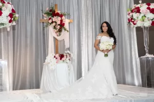 Professional Photographer A bride in an off-shoulder white gown smiles, bouquet in hand, standing on a stage with a floral-decorated cross and vibrant blooms. Captured by a skilled photographer, the scene glows against a shimmering silver curtain backdrop. www.maranonimagery.com