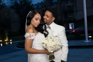 Professional Photographer A bride in an off-shoulder lace wedding dress holds a bouquet of white flowers, while the groom in a white suit smiles lovingly at her outdoors in the evening, beautifully captured by a skilled photographer. www.maranonimagery.com
