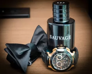 Professional Photographer A black bottle labeled Sauvage sits on a wooden surface, beside a black bow tie and a gold-accented wristwatch—perfect accessories for headshots capturing the elegance of men's fashion and grooming. www.maranonimagery.com