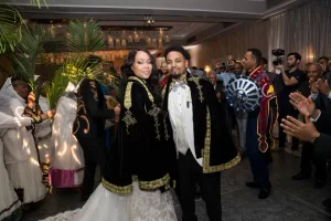 Professional Photographer A photographer captures a bride and groom wearing traditional black and gold habesha ceremonial capes as they smile at their wedding, surrounded by applauding guests in festive attire inside a decorated hall. www.maranonimagery.com
