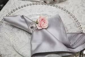 Professional Photographer A silver satin napkin is neatly folded and held with a jeweled napkin ring and a small pink rose, placed on a clear glass plate atop a white lace tablecloth—perfect for elegant wedding photography. www.maranonimagery.com