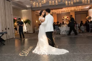 Professional Photographer A bride and groom share a first dance in an elegant ballroom, surrounded by guests at decorated tables. The bride wears a white gown, the groom a white jacket. Wedding photography beautifully captures Dina illuminated on the dance floor. www.maranonimagery.com