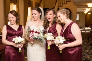 Professional Photographer Four women stand together indoors, smiling and holding bouquets. Three wear matching sleeveless burgundy dresses, while the bride in a white lace gown holds a larger bouquet. Captured by a skilled photographer, they appear to be celebrating a wedding. www.maranonimagery.com