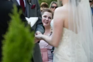 Professional Photographer A woman in a pink dress smiles warmly while watching a bride and groom hold hands during an outdoor wedding ceremony, captured beautifully by the photographer. The bride is wearing a veil and lace dress, seen from behind. www.maranonimagery.com