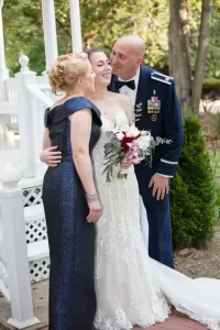 Professional Photographer A bride in a lace wedding dress smiles, holding a bouquet, as she is embraced by a woman in a dark blue gown and a man in a formal military uniform, all posing outdoors by white steps for timeless portraits captured by the photographer. www.maranonimagery.com