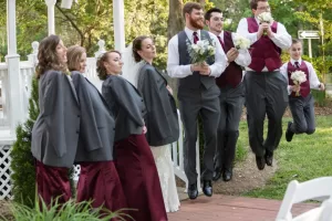 Professional Photographer A bridal party poses playfully outdoors; the Photographer captures bridesmaids in oversized suit jackets and groomsmen in maroon vests, all jumping and holding bouquets, smiling and having fun. www.maranonimagery.com