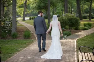 Professional Photographer A bride and groom walk hand in hand down a paved path in a park, the bride in a white gown with a veil holding a bouquet, and the groom in a gray suit. Wedding photography captures them beautifully among the trees and greenery. www.maranonimagery.com