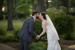 Professional Photographer A bride in a lace wedding dress and veil kisses a groom in a gray suit as they hold hands outdoors in a green, wooded area—an enchanting moment captured through expert wedding photography. www.maranonimagery.com
